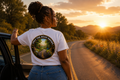 Person standing by a car on a road at sunset, wearing a white t-shirt with a graphic design.